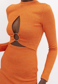 Robe en maille orange avec un col montant, des manches longues et une découpe en forme de goutte ornée d’un anneau argenté. Présente un motif géométrique sur toute la surface.