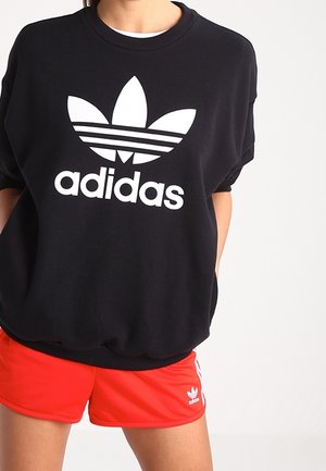 Osoba nosząca czarną bluzę Adidas z białym logo oraz czerwone spodenki Adidas z białymi paskami i małym logo na lewej nodze.