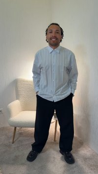 Άνδρας με κοντά dreadlocks χαμογελάει ενώ στέκεται με τα χέρια στις τσέπες, φορώντας ένα ανοιχτό ριγέ πουκάμισο, μαύρο παντελόνι και μαύρα παπούτσια μέσα σε εσωτερικό χώρο.