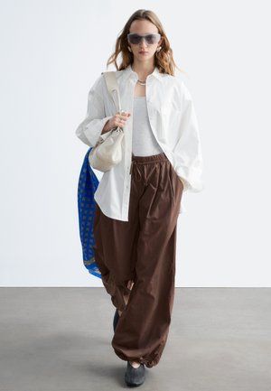 Mujer con camisa blanca extragrande, camiseta sin mangas gris, pantalones anchos marrones, zapatos negros, gafas de sol grandes, llevando dos bolsos, caminando en interiores.