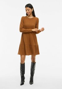 Femme ajustant la manche d'une robe longue à manches ribbées marron, portant des bottes noires à talons jusqu'au genou, se tenant contre un fond blanc uni.