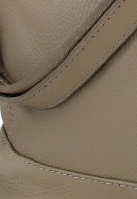 Handtas van lichtbruin leer met een getextureerd oppervlak, met opvallende stiksels en een gebogen ontwerp. Voorzien van een overslagklep en een verstelbare band.