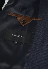 Innenansicht eines marineblauen Sakkos, mit einem braunen Akzentknopf, einem Stoffetikett mit "Matinique" und einem strukturierten marineblauen Stoffmuster.