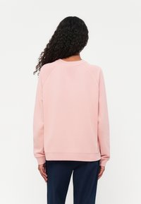 Sweatshirt rosa chiaro con maniche raglan, scollo e polsini a costine, e una trama liscia. Abbinato a pantaloni blu scuro.