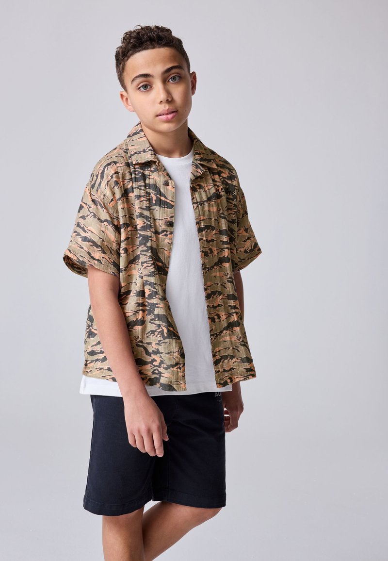 Korte mouwen, shirt met camouflagepatroon in beige, groen en zwart, gedragen over een wit T-shirt, gecombineerd met zwarte shorts, met een ontspannen pasvorm.