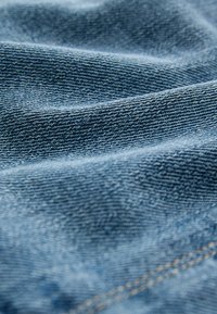 Denim-kangas vaaleansinisessä värissä, jossa on sileä pinta, esittelee vaakasuuntaisia kudontamalleja ja hienovaraisia haalistumia. Kultaiset tikkauslangat näkyvät saumoissa.