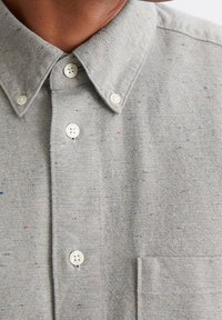 Nahaufnahme eines grauen Hemdes mit weißen Knöpfen und einer Brusttasche, getragen von einer Person mit sichtbarem Hals und Kinn.