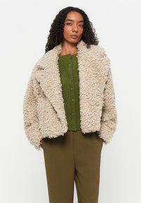 Beige fluffig shearlingjacka med överdimensionerade slag, bärs över en grön ribbad knapplös topp, tillsammans med olivfärgade byxor.
