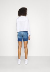Camisa blanca corta con botones y mangas largas, combinada con pantalones cortos de mezclilla azules. Modelo usa zapatillas blancas, mostrando un look casual.