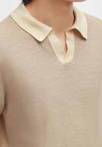 Beige stickad polo tröja med krage och texturerad väv, med en v-springa framtill och kontrasterande ljusa krage detaljer.