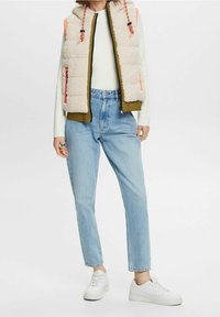 Lichte, mouwloze bodywarmer in beige fleece met olijfgroene accenten. Voorzien van een hoge kraag, oranje details en ritssluitingen aan de zakken. Gecombineerd met een blauwe spijkerbroek.