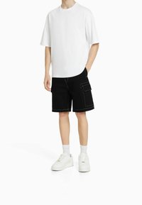 Valkoinen puuvillainen ylisuurikokoinen t-paita yhdistettynä mustiin cargo-shorteihin, joissa on etu- ja sivutaskut sekä kontrastivalkoiset tikkausompeleet. Yhdistetty valkoisiin lenkkareihin.