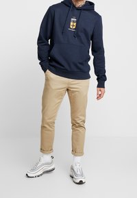 Sudadera navy con un diseño gráfico de batería en amarillo, combinada con chinos beige y zapatillas grises. Presenta una capucha con cordón y puños acanalados.