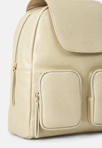 Sac à dos en similicuir crème avec surface texturée, deux poches avant zippées, fermeture à rabat et détails en métal doré.