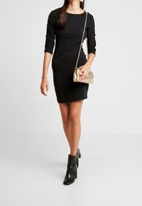 Musta, vartalonmyötäinen mekko, jossa on kolme neljäsosaa hihaa ja olkatekoja. Yhdistetty mustiin korollisiin nilkkureihin ja kultaan crossbody-laukku.