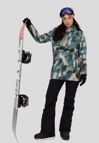 Žena v maskáčové bundě, černých kalhotách, rukavicích a lyžařských brýlích drží snowboard vztyčený proti světlému šedému pozadí.