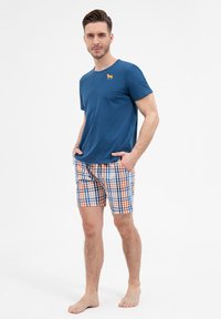 Niebieski t-shirt z krótkim rękawem z małym pomarańczowym logo psa, zestawiony z kratkowanymi szortami w niebieskim, pomarańczowym i białym kolorze. Model stoi boso.