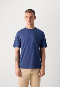 T-shirtissä on lyhyet hihat ja se on tummansinisestä puuvillasta. Vasemmassa rinnassa on pieni musta graafinen printti. Klassinen pyöreä kaula-aukko.