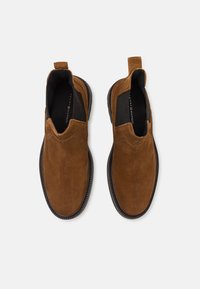 Pantofi slip-on din suede maro, cu o textură netedă, panouri elastice pe laterale și o talpă din cauciuc negru. Prezintă un toc mic și detalii cu cusătură.