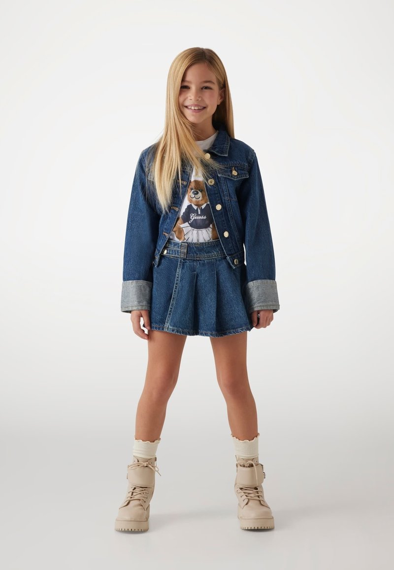 Jeansjakke med lys grå manchetter, grafisk T-shirt med et bjørnemotiv, denimnederdel og beige snørebåndsstøvler med cremefarvede strømper.