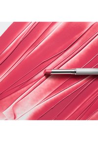 Un produit pour les lèvres rose repose sur une surface rose texturée et brillante avec des lignes ondulées, mettant en valeur sa consistance lisse et crémeuse. Accent métallique argenté sur l'applicateur.