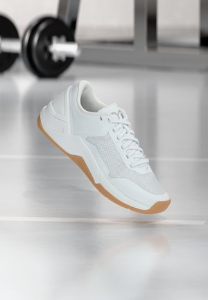 Fehér sportcipő gumi talppal, amely egy tükröződő edzőtermi padló felett lebeg, homályos súlyzó és állvány a háttérben.