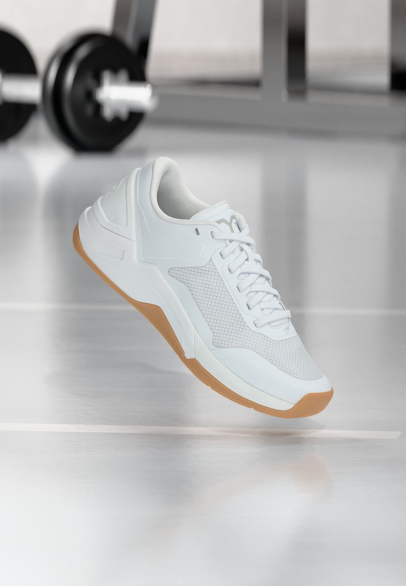 Άσπρο αθλητικό παπούτσι με λαστιχένια σόλα αιωρείται πάνω από αντανάκλαση γυμναστηριακού δαπέδου, θολό αλτήρα και ράφι στο φόντο.