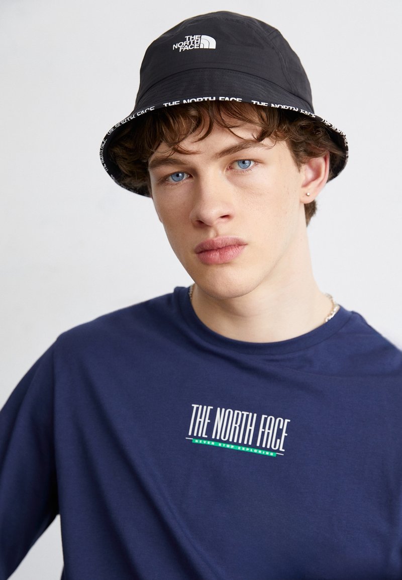 Jeune homme aux yeux bleus portant un chapeau bob noir The North Face et une chemise bleu marine avec le logo The North Face sur la poitrine.