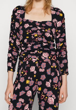 Femme portant un haut noir froncé et un pantalon assorti aux motifs floraux roses, jaunes et bleus, avec de longues manches bouffantes.