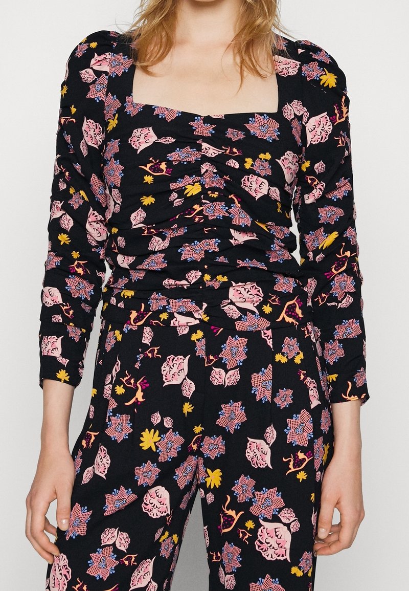 Femme portant un haut noir froncé et un pantalon assorti aux motifs floraux roses, jaunes et bleus, avec de longues manches bouffantes.