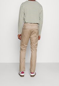 Person i beige byxor, vita sneakers med röda detaljer, och en långärmad vit och olivgrön randig tröja, som står mot en enfärgad bakgrund.