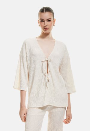 Blusa bianca in tessuto con scollo a V e dettagli con nodo sul davanti. Presenta maniche ampie e una silhouette morbida. Abbinata a pantaloni coordinati.