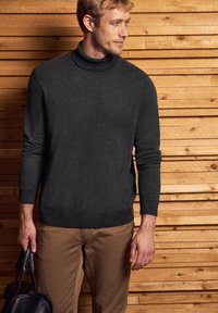 Suéter de cuello alto gris oscuro en un tejido texturizado, combinado con pantalones marrón claro. La figura sostiene un bolso negro y está de pie frente a tablas de madera.