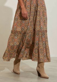 Dlouhé šaty s vzorovaným designem a vrstveným spodním lemem, které se vyznačují zemskými tóny a složitými vzory. Doplněné o světle hnědé kotníčkové boty.