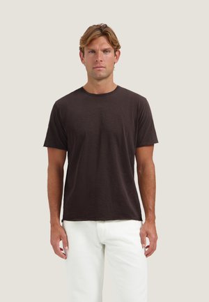 Man met lichtbruin haar, gekleed in een effen donkerbruine korte mouwen T-shirt en een witte broek, staand voor een neutrale achtergrond.