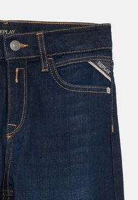 Blugi din denim albastru închis cu un design ajustat, cusături portocalii în contrast, închidere cu nasture și o etichetă mică a mărcii pe buzunar.