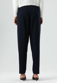 Marineblauwe, op maat gemaakte broek met een ontspannen pasvorm en geplooide voorkant, gecombineerd met lichtgekleurde schoenen, met een gladde stoftextuur.