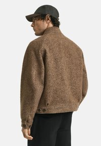 Bruine herringbone-textuur jasje met een hoge kraag, knoopdetails bij de manchet en een relaxte pasvorm, gecombineerd met een donkere pet.