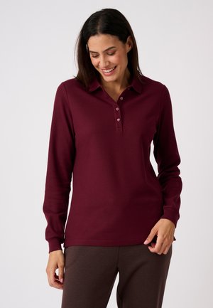 Femme souriante regardant vers le bas, portant un polo bordeaux à manches longues et un pantalon marron foncé sur un fond clair uni.
