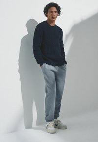 Mornarica pleteni pulover s kablasta uzorka, uparen sa sivim trenirkom i bijelim tenisicama. Model stoji nasuprot jednostavne pozadine, bacajući sjenu.