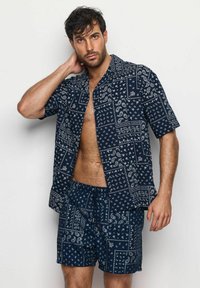 Muž s tmavými vlasy, oblečený v krátkém rukávu košili s námořnickým modrým vzorem bandany a odpovídajících šortkách, pózuje s jednou rukou za krkem.