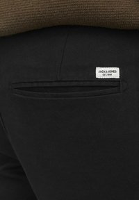 Black trousers featuring a back pocket with a narrow slit and a sewn-in label with "JACK & JONES EST. 1990" in white. Soft, textured fabric.