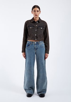 Frau steht nach vorne gerichtet, trägt eine schwarze, kurze Jeansjacke, weite blaue Jeans und schwarze Schuhe vor hellem Hintergrund.