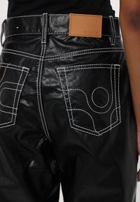 Pantaloni din piele negri cu cusături albe și două buzunare posterioare cu un design curbat distinct. O etichetă de marcă din piele bej este vizibilă pe talie.
