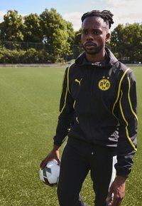 Chaqueta deportiva negra con acentos y logotipo amarillos, combinada con pantalones negros, sosteniendo un balón de fútbol, en un campo de césped artificial verde.