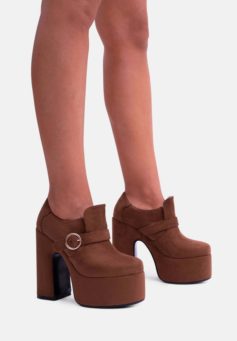 Tacco a piattaforma in suede marrone con un tacco spesso e un elegante dettaglio in fibbia metallica circolare. Presentano una punta arrotondata e una texture liscia.