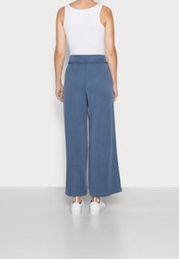 Breda byxor i mjukt blått tyg med en slät yta. Hög midja med en detaljerad knytning bak. Matchas med vita sneakers.