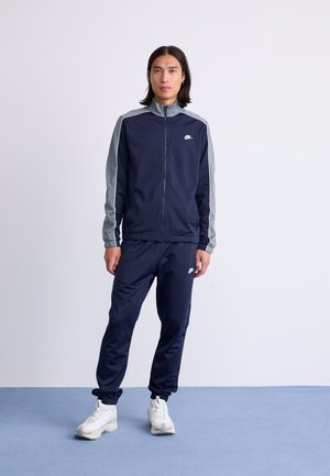 Survêtement bleu marine avec des accents gris aux épaules, veste zippée et pantalon à taille élastique, arborant le logo Nike blanc. Chaussures de sport blanches.