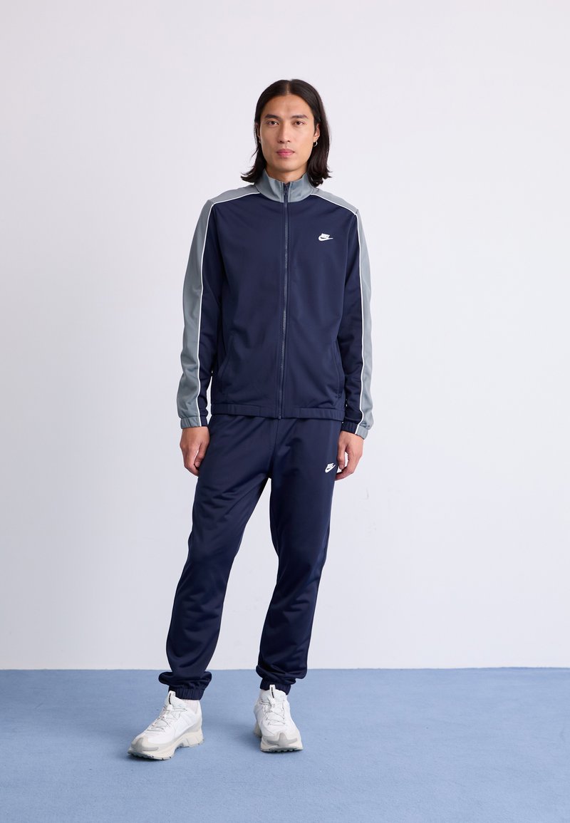Navy tracksuit with gray shoulder accents, zip-up jacket and elastic waistband pants, featuring white Nike logo. White athletic shoes.