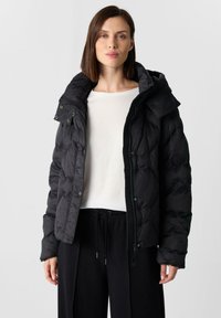 Kobieta w czarnej pikowanej kurtce z wysokim kołnierzem, białej bluzce z długim rękawem i czarnych spodniach ze sznurkiem, stoi na neutralnym tle.